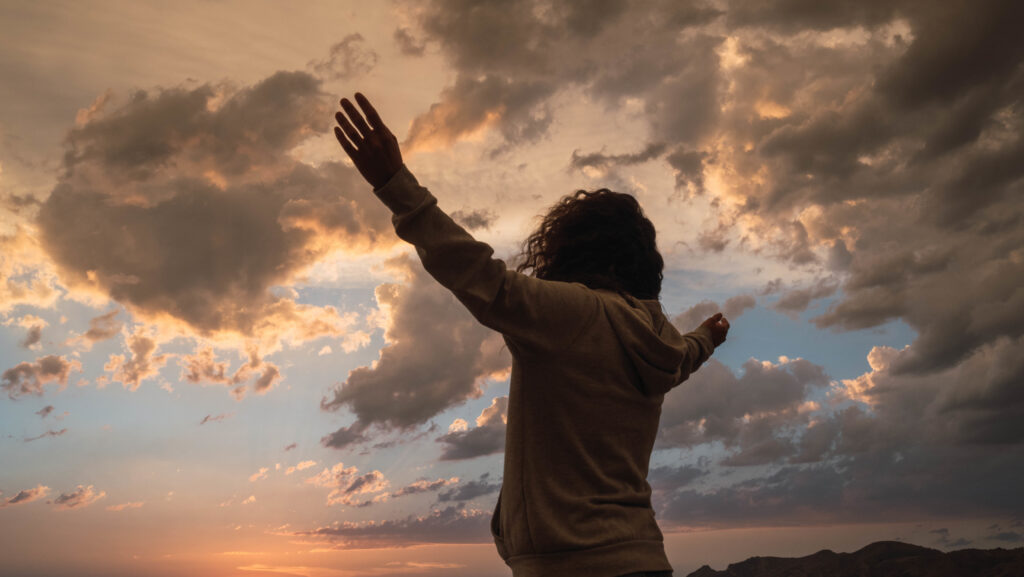 A back view of a female standing under the cloudy sunset sky with hands up